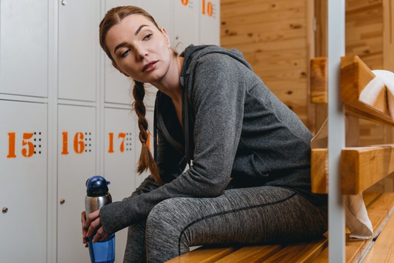 Woman in sportive attire sitting on bench in locker room with water bottle and looking aside