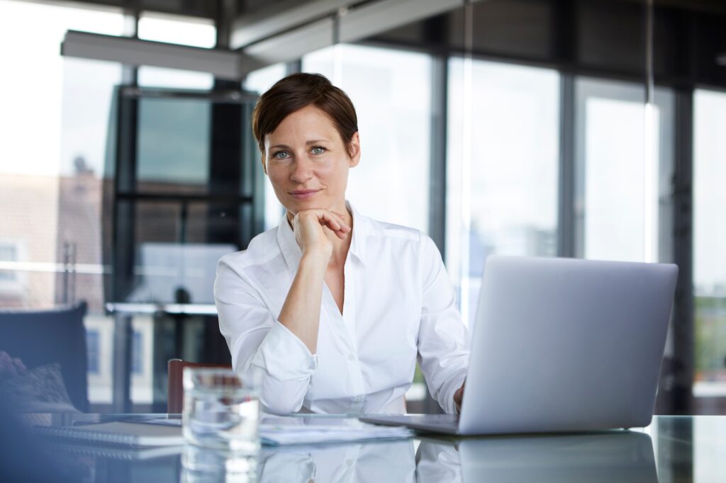 Portrait of confident businesswoman sitting at glass table in office with laptop